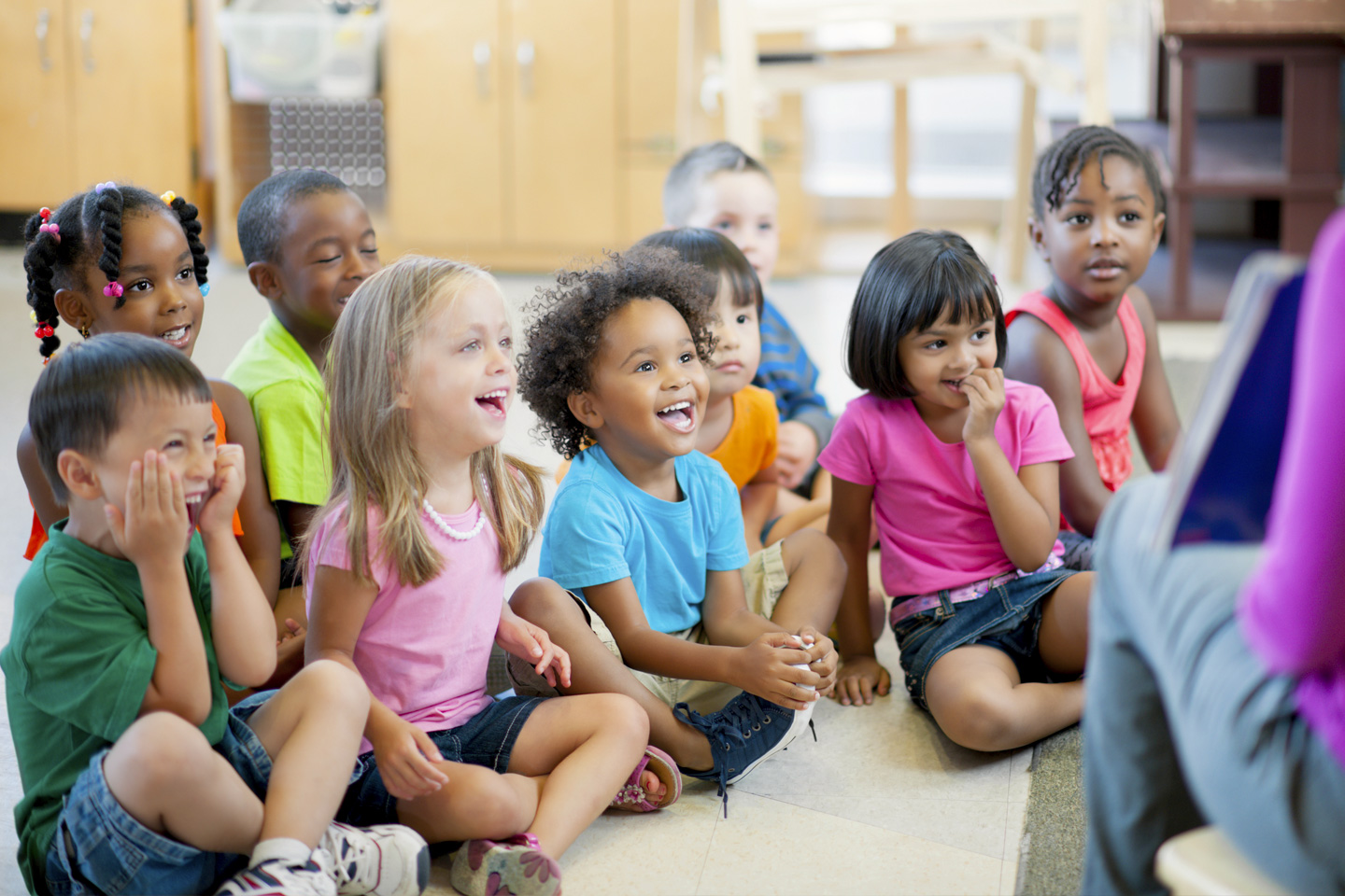 L'image montre un groupe d'enfants assis en tailleur sur le sol, visiblement captivés par une personne, possiblement un enseignant, qui se trouve devant eux et lit un livre. Les enfants, de différentes origines ethniques, portent des vêtements colorés et affichent des expressions de joie, d'émerveillement et d'intérêt. L'arrière-plan suggère une salle de classe ou un espace d'apprentissage préscolaire.